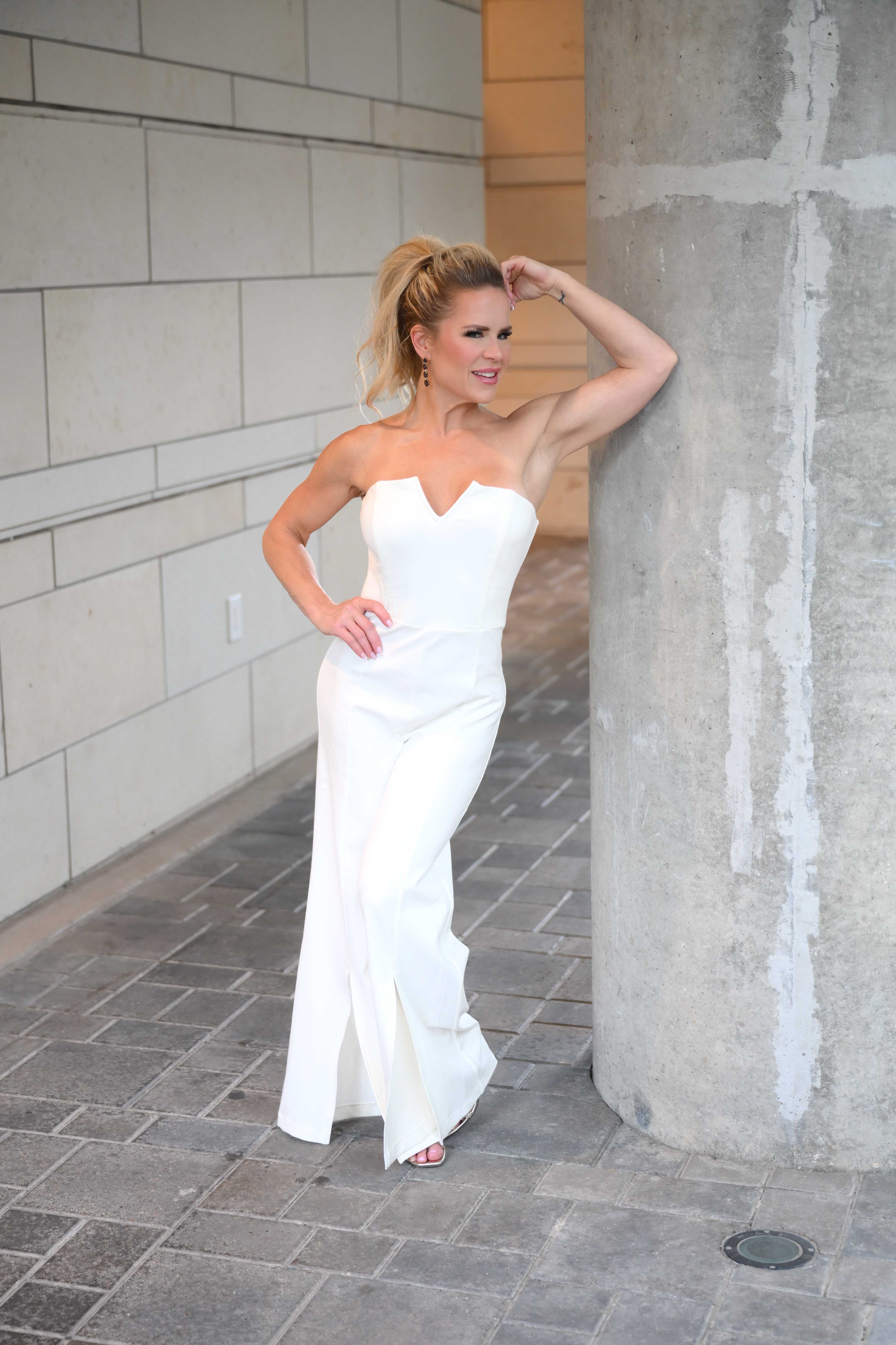 Janelle Alexis in white strapless jumpsuit leaning on architectural column — women empowerment speaker